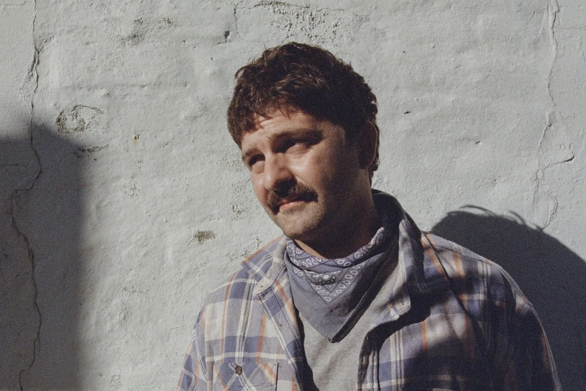 Australian indie-folk musician Adam Newling wears a plaid shirt and bandana, leaning against a white textured wall in sunlight, looking thoughtfully to the side.